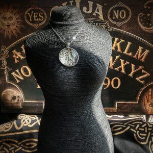 May include: A silver pendant necklace with a circular, clear resin pendant. The pendant contains a detailed, abstract design. The necklace is displayed on a black mannequin. The background features a Ouija board and other occult-themed elements.