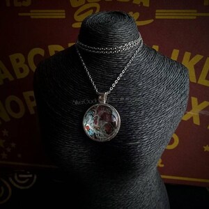 May include: A silver-toned pendant necklace with a round, glass-covered charm. The charm features a colorful, abstract design with red and blue accents. The necklace is displayed on a black mannequin.