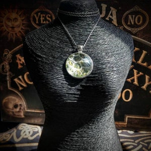May include: A silver-toned pendant necklace with a round, clear glass pendant. The pendant contains a green and white design. The necklace is displayed on a black mannequin. The background includes a Ouija board and other occult-themed items.