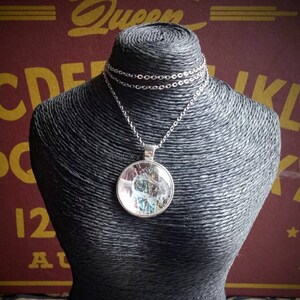 May include: A silver necklace with a round pendant on a black mannequin. The pendant has a clear glass or resin covering a colorful, abstract design. The necklace chain is delicate and hangs around the mannequin's neck.
