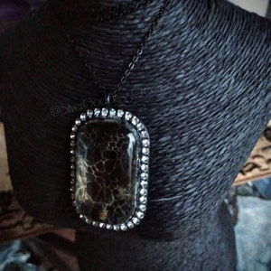 May include: A black necklace with a rectangular pendant. The pendant has a dark, patterned center and is framed by a row of small, clear stones. The necklace is displayed on a black bust.