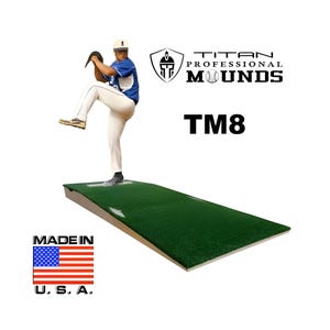 May include: A baseball pitcher in a blue and white uniform is captured mid-pitch on a green artificial turf pitching mound. The mound is labeled "TM8" and the logo for "Titan Professional Mounds" is visible. The words "Made in U.S.A." are displayed with the American flag.