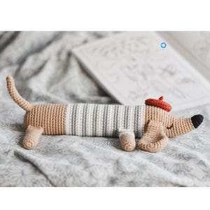 May include: Crocheted dachshund dog toy in beige, gray, and white. The dog wears a red beret and has a black nose. The toy is lying on a patterned blanket with a book in the background.