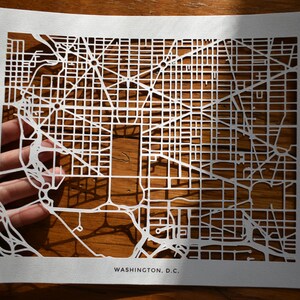 May include: A white paper map of Washington, D.C. with the city streets cut out. The map is on a wooden surface.