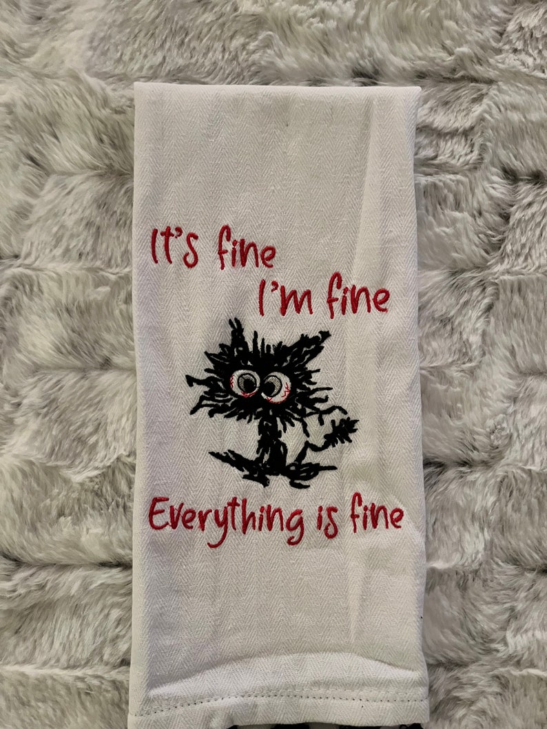 May include: White kitchen towel with red embroidered text that reads "It's fine I'm fine Everything is fine" and a black embroidered cartoon cat with big eyes.