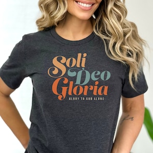 Op de afbeelding: Een donker grijs t-shirt met een retro-stijl grafische afbeelding met de tekst "Soli Deo Gloria" en "Glory to God Alone".