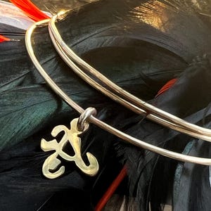 Puede incluir: Un brazalete de metal en tono dorado con acabado pulido y un dije colgante con forma de letra A estilizada. El brazalete está compuesto por dos bandas circulares finas y lisas que se superponen para formar un diseño de bucle continuo.