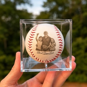 May include: A white baseball with red stitching encased in a clear square display box. The ball features a laser-engraved illustration of an adult and two children, with the text "Best Papa Ever" printed in a cursive font below the image.