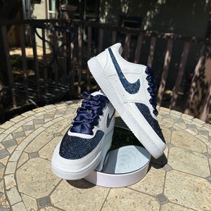 May include: White leather sneakers with a navy blue swoosh and a navy blue satin ribbon. The sneakers are covered in rhinestones.