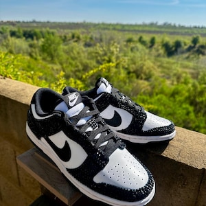 K&ouml;nnte beinhalten: Ein Paar schwarz-wei&szlig;e Nike Dunk Low Sneaker, verziert mit glitzernden schwarzen Akzenten. Die Schuhe haben eine wei&szlig;e Basis mit schwarzen Overlays, einen schwarzen Nike Swoosh und schwarze Schn&uuml;rsenkel. Die Sneaker werden im Freien vor dem Hintergrund von Gr&uuml;n und blauem Himmel pr&auml;sentiert.