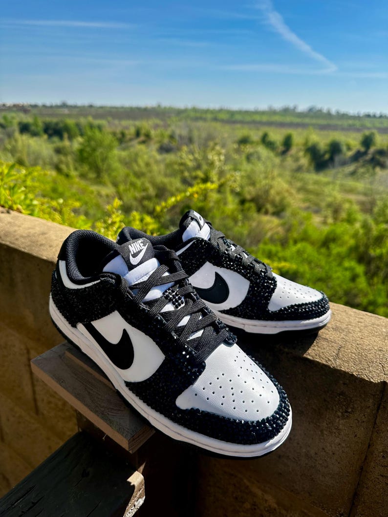 K&ouml;nnte beinhalten: Ein Paar schwarz-wei&szlig;e Nike Dunk Low Sneaker, verziert mit glitzernden schwarzen Akzenten. Die Schuhe haben eine wei&szlig;e Basis mit schwarzen Overlays, einen schwarzen Nike Swoosh und schwarze Schn&uuml;rsenkel. Die Sneaker werden im Freien vor dem Hintergrund von Gr&uuml;n und blauem Himmel pr&auml;sentiert.