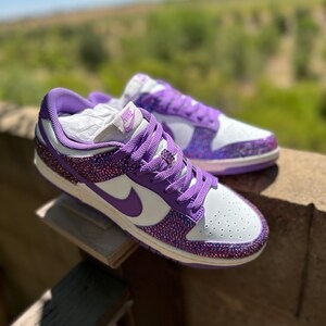 K&ouml;nnte beinhalten: Paar wei&szlig;-lila Turnschuhe mit glitzerndem Design. Die Schuhe haben lila Schn&uuml;rsenkel, einen lila Nike-Swoosh und eine wei&szlig;e Zehenbox. Die Turnschuhe werden im Freien pr&auml;sentiert.