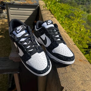 K&ouml;nnte beinhalten: Paar wei&szlig;-schwarze Nike Dunk Low Sneaker. Die Sneaker haben eine wei&szlig;e Basis mit schwarzen Akzenten, einschlie&szlig;lich des Nike-Swoosh und der Schn&uuml;rsenkel. Zehenbereich und Kanten sind mit schwarzen Strasssteinen verziert. Das Nike-Logo ist auf der Zunge sichtbar.