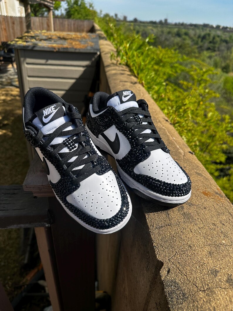 K&ouml;nnte beinhalten: Paar wei&szlig;-schwarze Nike Dunk Low Sneaker. Die Sneaker haben eine wei&szlig;e Basis mit schwarzen Akzenten, einschlie&szlig;lich des Nike-Swoosh und der Schn&uuml;rsenkel. Zehenbereich und Kanten sind mit schwarzen Strasssteinen verziert. Das Nike-Logo ist auf der Zunge sichtbar.