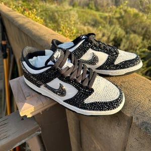 K&ouml;nnte beinhalten: Schwarze und wei&szlig;e Nike Dunk Low Sneaker, verziert mit glitzernden Kristallen. Die Schuhe haben ein wei&szlig;es Swoosh-Logo, schwarze Schn&uuml;rsenkel und eine wei&szlig;e Zwischensohle. Die Sneaker werden im Freien gezeigt.