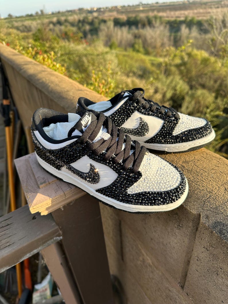 K&ouml;nnte beinhalten: Schwarze und wei&szlig;e Nike Dunk Low Sneaker, verziert mit glitzernden Kristallen. Die Schuhe haben ein wei&szlig;es Swoosh-Logo, schwarze Schn&uuml;rsenkel und eine wei&szlig;e Zwischensohle. Die Sneaker werden im Freien gezeigt.