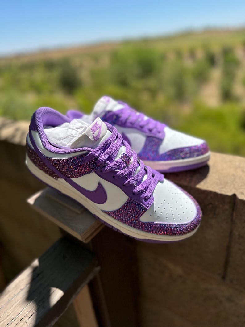 K&ouml;nnte beinhalten: Ein Paar wei&szlig;-lila Sneaker mit glitzerndem Design. Die Schuhe haben wei&szlig;e Zehenboxen und Seitenteile, mit einem lila Nike-Swoosh und Schn&uuml;rsenkeln. Ferse und Zehen sind mit schimmerndem Glitzer bedeckt.