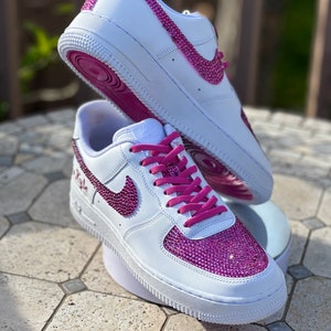 May include: White leather sneakers with pink laces and a pink rhinestone design on the swoosh and toe. The sneakers have a white sole and a pink outsole.