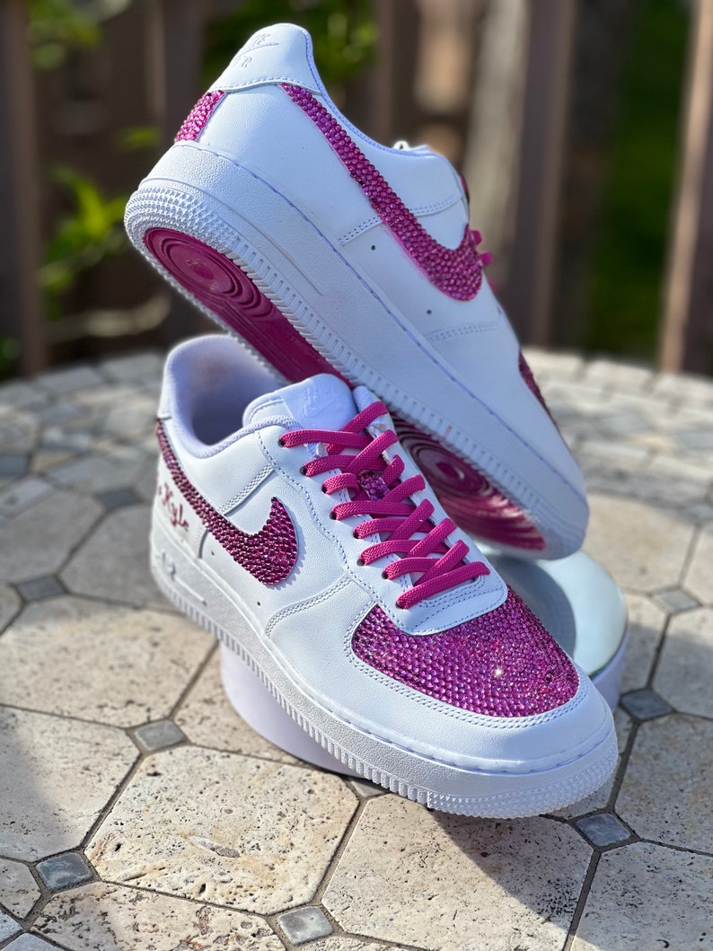 May include: White leather sneakers with pink laces and a pink rhinestone design on the swoosh and toe. The sneakers have a white sole and a pink outsole.