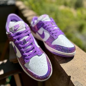 K&ouml;nnte beinhalten: Ein Paar wei&szlig;-lila Turnschuhe mit glitzerndem, geschm&uuml;cktem Design. Die Schuhe zeigen das Nike-Logo auf der Zunge und einen lila Swoosh. Der Zehenbereich ist wei&szlig; mit kleinen Perforationen.