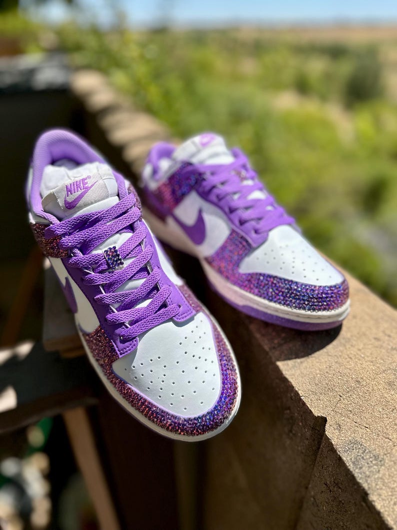 K&ouml;nnte beinhalten: Ein Paar wei&szlig;-lila Turnschuhe mit glitzerndem, geschm&uuml;cktem Design. Die Schuhe zeigen das Nike-Logo auf der Zunge und einen lila Swoosh. Der Zehenbereich ist wei&szlig; mit kleinen Perforationen.