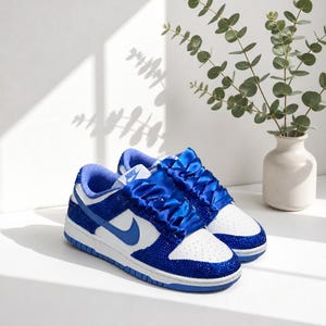 May include: A pair of white and royal blue sneakers featuring a white leather base with royal blue overlays covered in small sparkling crystals. The shoes have wide royal blue satin ribbon laces and a blue Nike swoosh on the side panel.