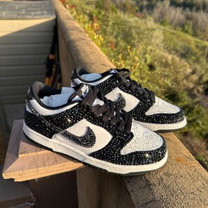 K&ouml;nnte beinhalten: Paar schwarze und wei&szlig;e Nike Dunk Low Sneaker, verziert mit glitzernden schwarzen und klaren Kristallen. Die Schuhe haben ein wei&szlig;es Swoosh-Logo und schwarze Schn&uuml;rsenkel, vor einem nat&uuml;rlichen Hintergrund im Freien.