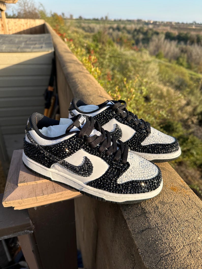 K&ouml;nnte beinhalten: Paar schwarze und wei&szlig;e Nike Dunk Low Sneaker, verziert mit glitzernden schwarzen und klaren Kristallen. Die Schuhe haben ein wei&szlig;es Swoosh-Logo und schwarze Schn&uuml;rsenkel, vor einem nat&uuml;rlichen Hintergrund im Freien.