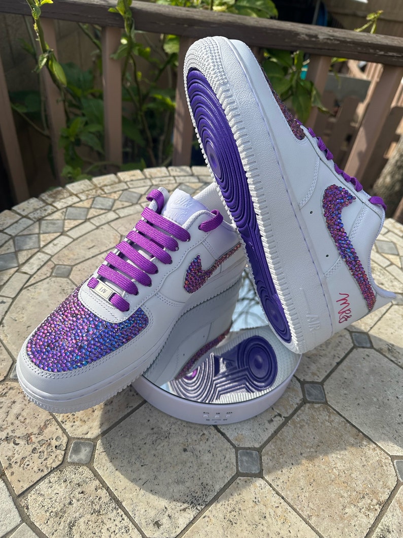 May include: White leather sneakers with purple laces and a purple trim. The sneakers are covered in iridescent rhinestones and have the word "MRS" written on the side.