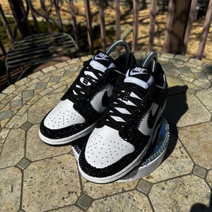 K&ouml;nnte beinhalten: Ein Paar schwarz-wei&szlig;e Nike Dunk Low Sneaker mit Kristallverzierungen. Die Schuhe haben eine wei&szlig;e Basis mit schwarzen Akzenten, einschlie&szlig;lich des Nike-Swoosh und der Schn&uuml;rsenkel. Der Zehenbereich ist perforiert.