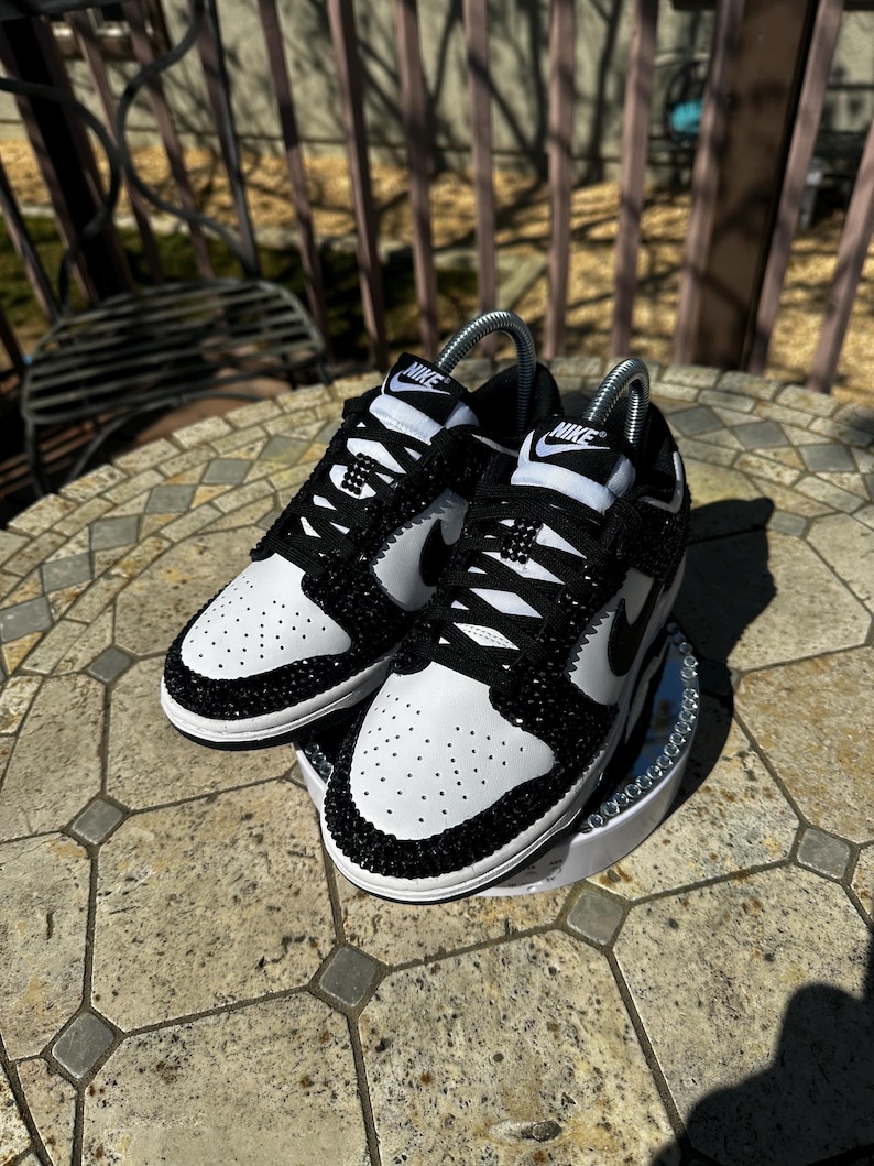 K&ouml;nnte beinhalten: Ein Paar schwarz-wei&szlig;e Nike Dunk Low Sneaker mit Kristallverzierungen. Die Schuhe haben eine wei&szlig;e Basis mit schwarzen Akzenten, einschlie&szlig;lich des Nike-Swoosh und der Schn&uuml;rsenkel. Der Zehenbereich ist perforiert.