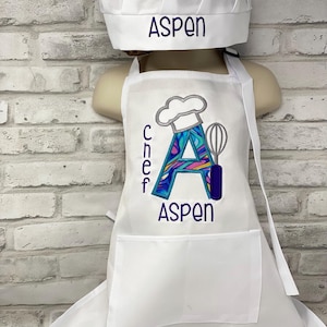 K&ouml;nnte beinhalten: Wei&szlig;es Kochm&uuml;tzen- und Sch&uuml;rzen-Set. Auf der M&uuml;tze ist "ASPEN" in Lila gestickt. Die Sch&uuml;rze zeigt ein farbenfrohes "A" mit Kochm&uuml;tzen- und Schneebesen-Design und den Namen "ASPEN" in Lila.