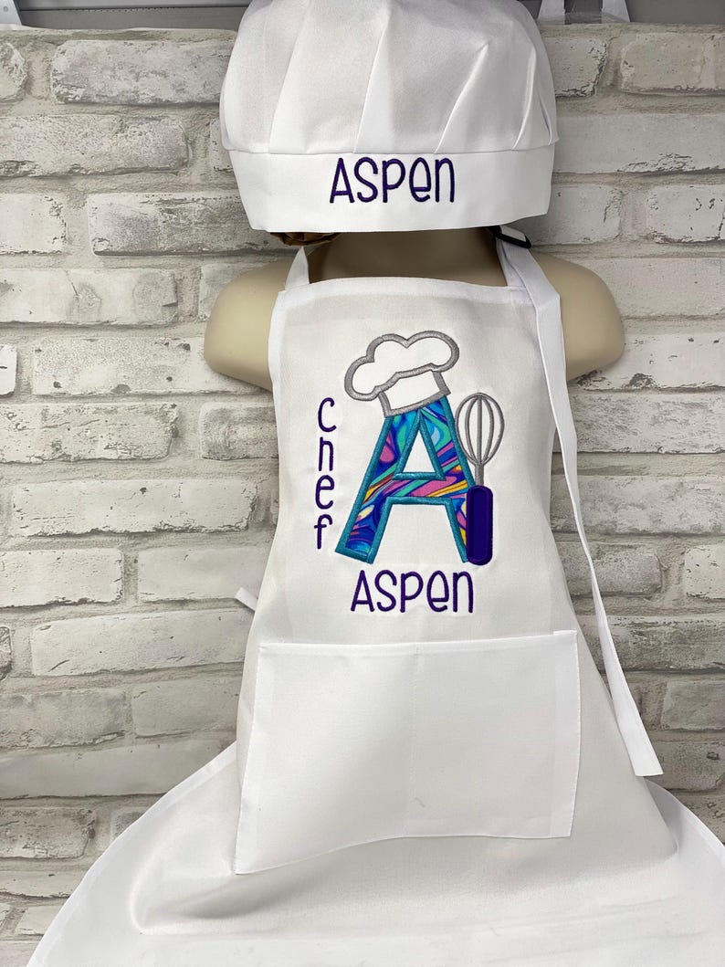 K&ouml;nnte beinhalten: Wei&szlig;es Kochm&uuml;tzen- und Sch&uuml;rzen-Set. Auf der M&uuml;tze ist "ASPEN" in Lila gestickt. Die Sch&uuml;rze zeigt ein farbenfrohes "A" mit Kochm&uuml;tzen- und Schneebesen-Design und den Namen "ASPEN" in Lila.