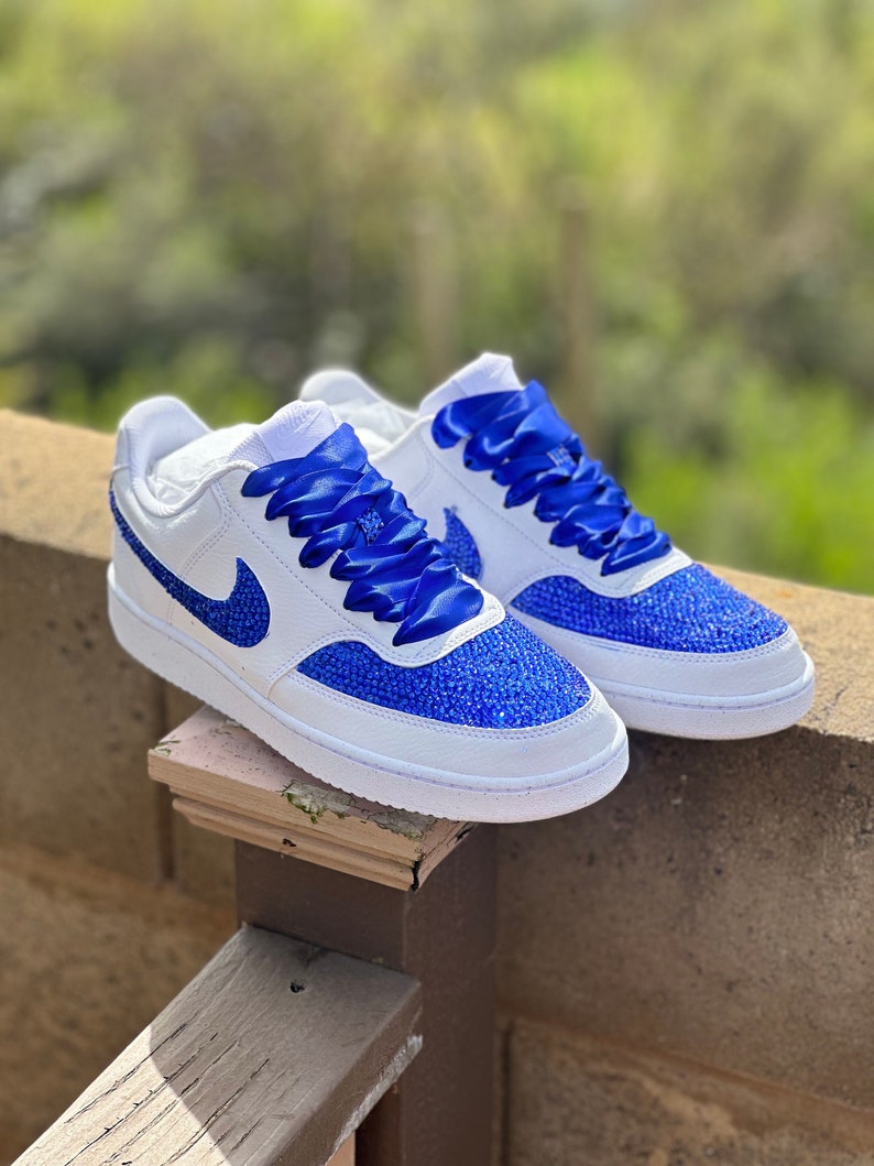 May include: White leather sneakers with blue ribbon laces and blue rhinestone detailing.