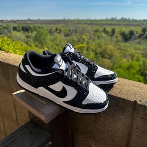 K&ouml;nnte beinhalten: Ein Paar schwarz-wei&szlig;e Sneaker mit einem Schmuckdesign. Die Schuhe haben einen schwarzen Nike-Swoosh und schwarze Schn&uuml;rsenkel. Die Sneaker werden auf einer Holzoberfl&auml;che mit nat&uuml;rlichem Hintergrund pr&auml;sentiert.