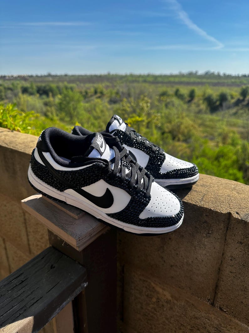 K&ouml;nnte beinhalten: Ein Paar schwarz-wei&szlig;e Sneaker mit einem Schmuckdesign. Die Schuhe haben einen schwarzen Nike-Swoosh und schwarze Schn&uuml;rsenkel. Die Sneaker werden auf einer Holzoberfl&auml;che mit nat&uuml;rlichem Hintergrund pr&auml;sentiert.