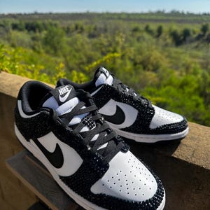 K&ouml;nnte beinhalten: Ein Paar schwarz-wei&szlig;e Nike Dunk Low Sneaker. Die Schuhe haben eine wei&szlig;e Basis mit schwarzen Akzenten, einschlie&szlig;lich des Nike-Swoosh und einem schwarzen, mit Juwelen besetzten Design um Zehen und Ferse. Die Schn&uuml;rsenkel sind schwarz.