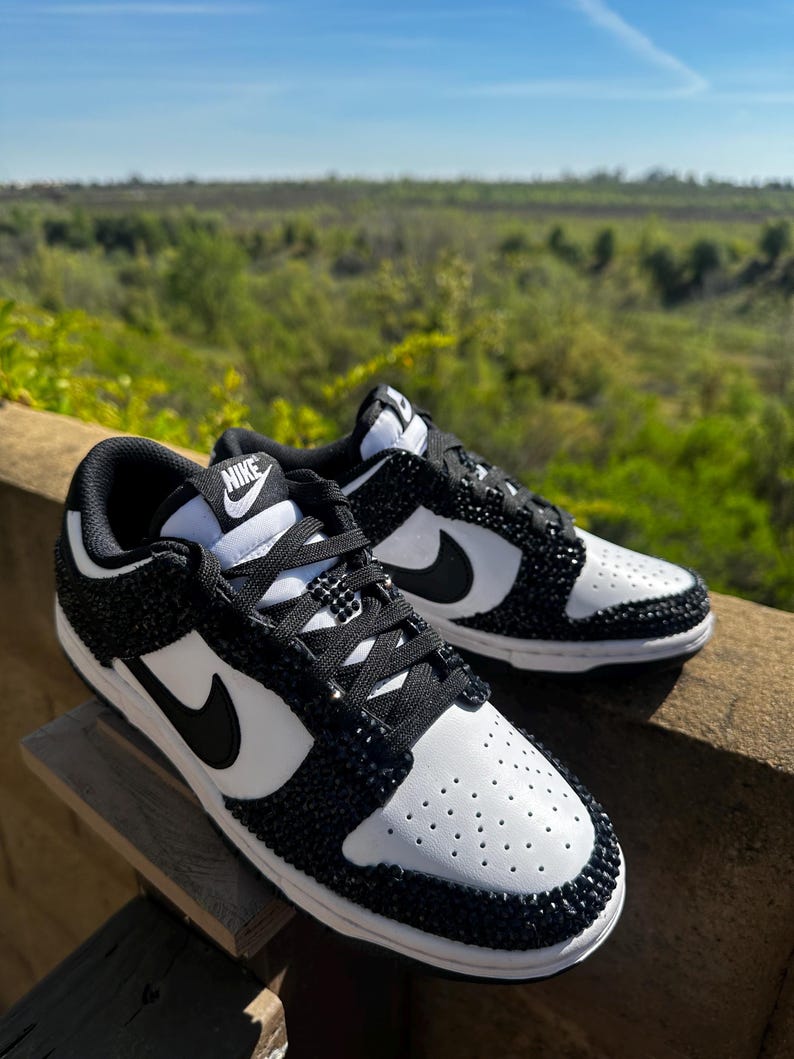 K&ouml;nnte beinhalten: Ein Paar schwarz-wei&szlig;e Nike Dunk Low Sneaker. Die Schuhe haben eine wei&szlig;e Basis mit schwarzen Akzenten, einschlie&szlig;lich des Nike-Swoosh und einem schwarzen, mit Juwelen besetzten Design um Zehen und Ferse. Die Schn&uuml;rsenkel sind schwarz.