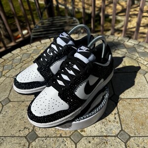 K&ouml;nnte beinhalten: Ein Paar schwarz-wei&szlig;e Nike Dunk Low Sneaker, verziert mit glitzernden schwarzen Kristallen. Die Schuhe haben eine wei&szlig;e Zehenbox und Seitenteile, mit einem schwarzen Nike Swoosh und Schn&uuml;rsenkeln. Die Sneaker werden auf einer wei&szlig;en Plattform pr&auml;sentiert.