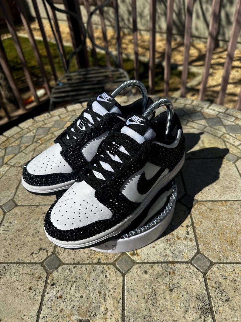 K&ouml;nnte beinhalten: Ein Paar schwarz-wei&szlig;e Nike Dunk Low Sneaker, verziert mit glitzernden schwarzen Kristallen. Die Schuhe haben eine wei&szlig;e Zehenbox und Seitenteile, mit einem schwarzen Nike Swoosh und Schn&uuml;rsenkeln. Die Sneaker werden auf einer wei&szlig;en Plattform pr&auml;sentiert.