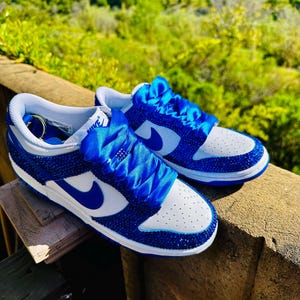 K&ouml;nnte beinhalten: Ein Paar blau-wei&szlig;e Sneaker mit einem Schmuckdesign. Die Sneaker haben eine wei&szlig;e Zehenbox, blaue Akzente und einen blauen Nike-Swoosh. Die Schn&uuml;rsenkel sind durch blaue Satinb&auml;nder ersetzt. Die Schuhe werden im Freien pr&auml;sentiert.