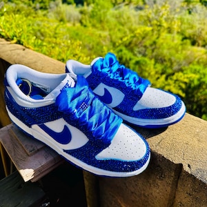 K&ouml;nnte beinhalten: Ein Paar blau-wei&szlig;e Sneaker mit einem Schmuckdesign. Die Sneaker haben eine wei&szlig;e Zehenbox, blaue Akzente und einen blauen Nike-Swoosh. Die Schn&uuml;rsenkel wurden durch blaue Satinb&auml;nder ersetzt. Die Schuhe werden im Freien pr&auml;sentiert.