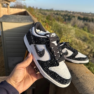 K&ouml;nnte beinhalten: Schwarz-wei&szlig;e Nike Dunk Low Sneaker mit Kristallverzierungen. Die Sneaker haben ein wei&szlig;es Swoosh-Logo, schwarze Schn&uuml;rsenkel und eine wei&szlig;e Zwischensohle. Die Schuhe werden im Freien gezeigt.