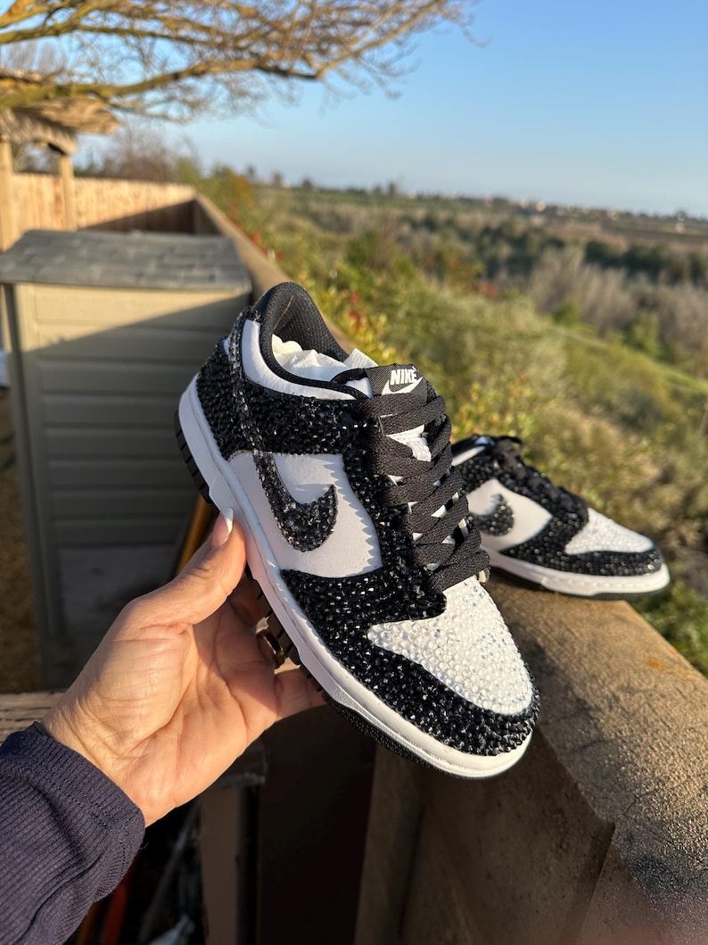 K&ouml;nnte beinhalten: Schwarz-wei&szlig;e Nike Dunk Low Sneaker mit Kristallverzierungen. Die Sneaker haben ein wei&szlig;es Swoosh-Logo, schwarze Schn&uuml;rsenkel und eine wei&szlig;e Zwischensohle. Die Schuhe werden im Freien gezeigt.