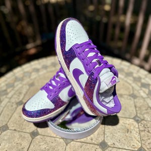 K&ouml;nnte beinhalten: Wei&szlig;e und lila Turnschuhe mit einem verzierten Design. Die Schuhe haben eine wei&szlig;e Basis mit lila Akzenten, einschlie&szlig;lich der Schn&uuml;rsenkel, des Nike-Swoosh und eines funkelnden, juwelenbesetzten &Uuml;berzugs auf Zehen und Ferse.