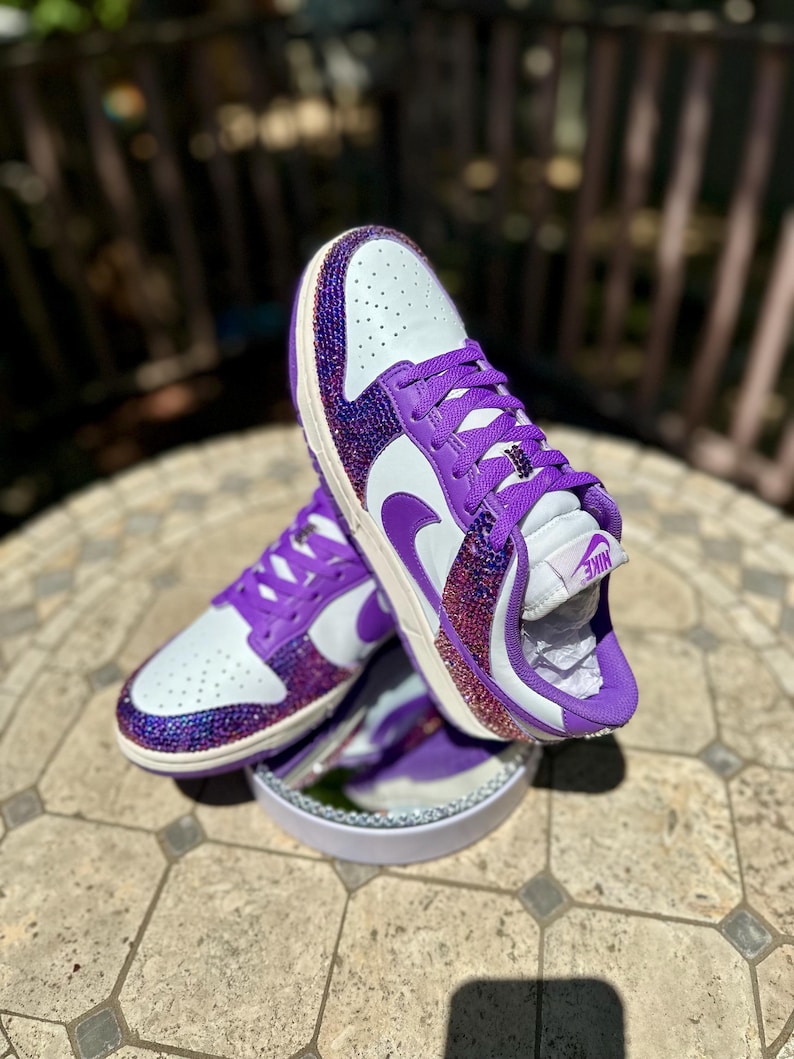 K&ouml;nnte beinhalten: Wei&szlig;e und lila Turnschuhe mit einem verzierten Design. Die Schuhe haben eine wei&szlig;e Basis mit lila Akzenten, einschlie&szlig;lich der Schn&uuml;rsenkel, des Nike-Swoosh und eines funkelnden, juwelenbesetzten &Uuml;berzugs auf Zehen und Ferse.