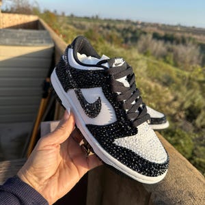 K&ouml;nnte beinhalten: Schwarz-wei&szlig;e Nike Dunk Low Sneaker mit Kristallverzierungen. Zehenbox und Seitenteile sind wei&szlig;, der Rest des Schuhs ist schwarz. Der Nike-Swoosh ist wei&szlig;. Schwarze Schn&uuml;rsenkel und eine wei&szlig;e Sohle.