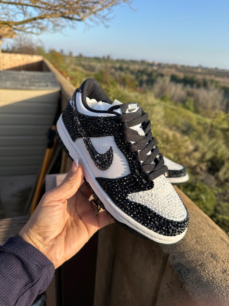 K&ouml;nnte beinhalten: Schwarz-wei&szlig;e Nike Dunk Low Sneaker mit Kristallverzierungen. Zehenbox und Seitenteile sind wei&szlig;, der Rest des Schuhs ist schwarz. Der Nike-Swoosh ist wei&szlig;. Schwarze Schn&uuml;rsenkel und eine wei&szlig;e Sohle.