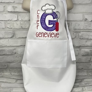 K&ouml;nnte beinhalten: Weisse Sch&uuml;rze mit grosser Vordertasche und verstellbarem Nackenband. Die Sch&uuml;rze tr&auml;gt den Namen "Genevieve" in Burgunderrot gestickt, zusammen mit dem Wort "Chef" und einem Kochm&uuml;tzen-Design. Der Buchstabe "G" ist in einem lila Design.