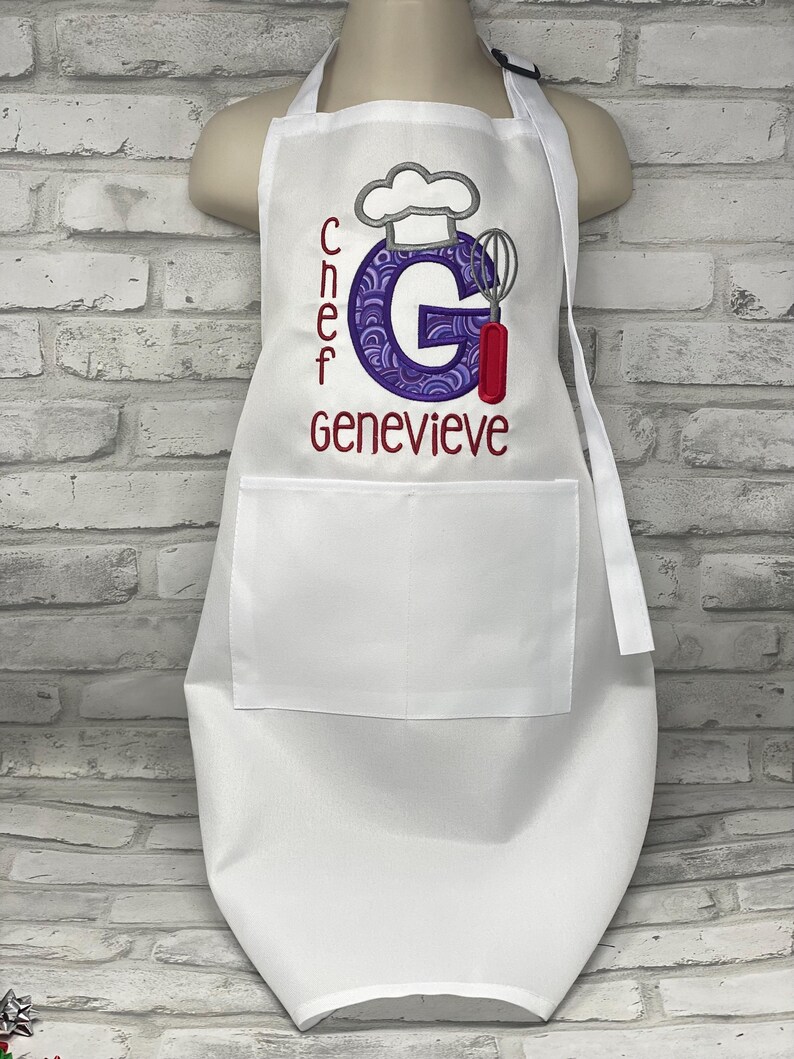 K&ouml;nnte beinhalten: Weisse Sch&uuml;rze mit grosser Vordertasche und verstellbarem Nackenband. Die Sch&uuml;rze tr&auml;gt den Namen "Genevieve" in Burgunderrot gestickt, zusammen mit dem Wort "Chef" und einem Kochm&uuml;tzen-Design. Der Buchstabe "G" ist in einem lila Design.